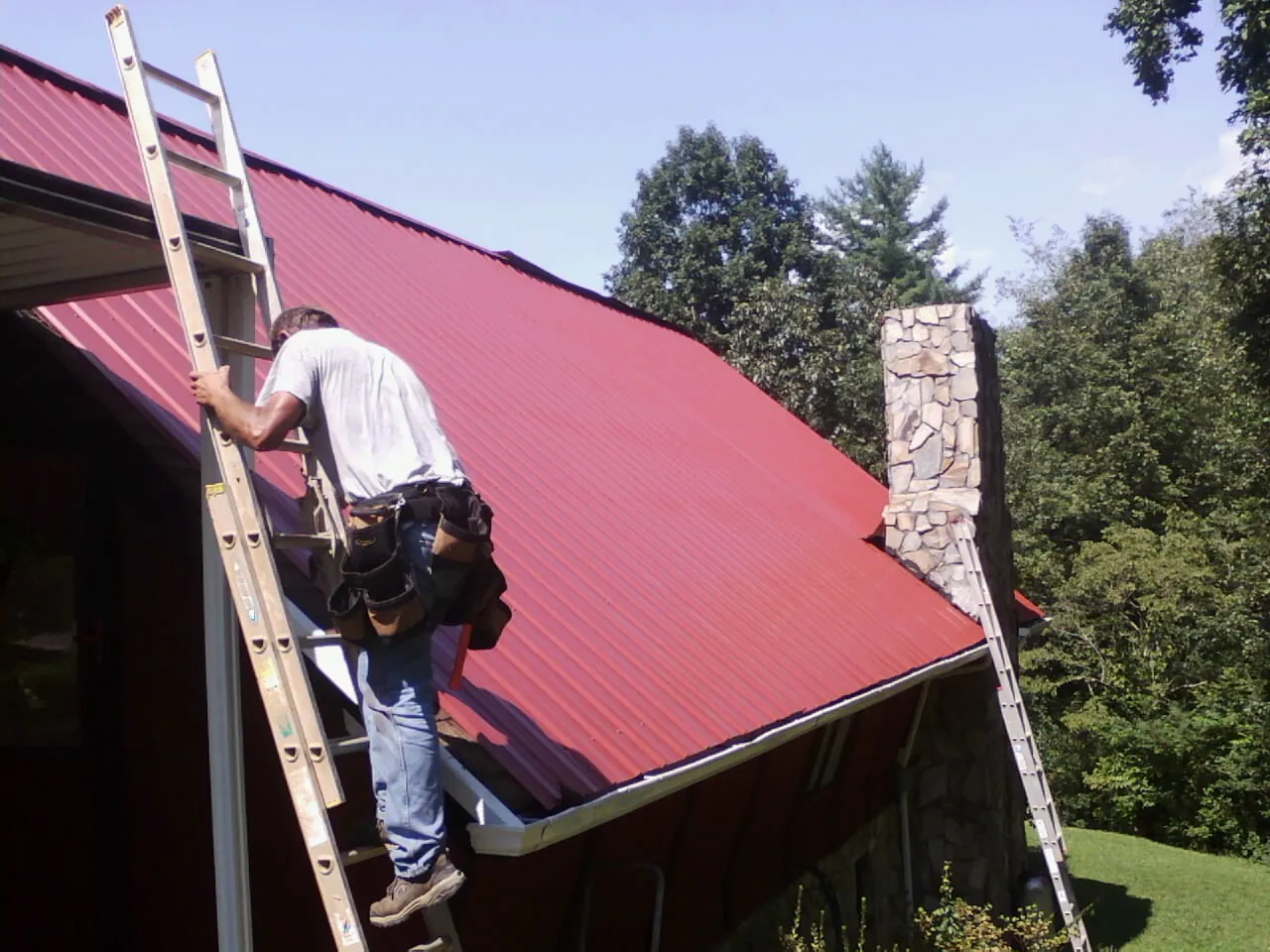 Roofer on a ladder beside a red metal roof and stone chimney — Soffit & Fascia Repair by Roofing Brothers in Velva
